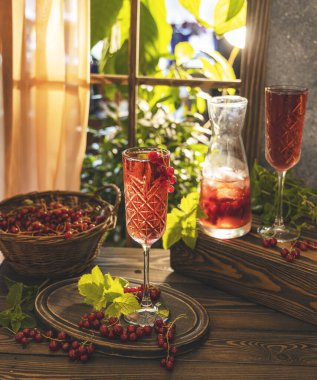 Pencerenin yanındaki masada iki bardak taze soğuk kuşüzümü kokteyli, güneşli arka plan ışığı. Koyu ahşap.