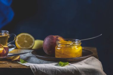 Taze ev yapımı şeftali reçeli kavanoza doluyor. Eski karanlık kırsal sihir tarzı. Güneşli bir ışık. Yanında birkaç taze meyve, limon ve nane var..