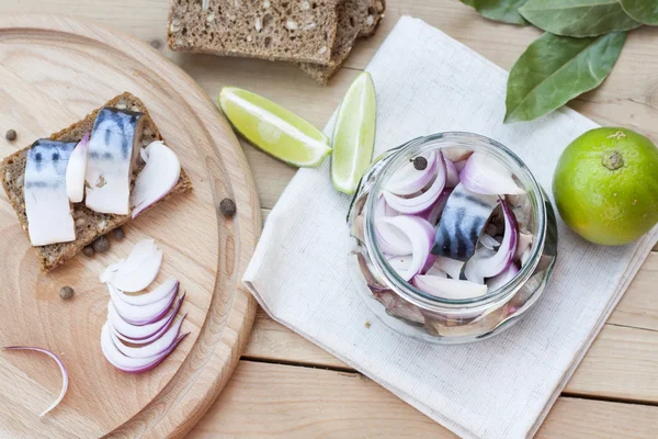 Slices of marinated mackerel with onion in a jar, lime, laurel and bread on wooden board — Stock Photo, Image