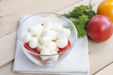 Mozzarella cheese in a glass bowl, tomatoes, sliced tomatoes and herbs on a wooden table, selective focus