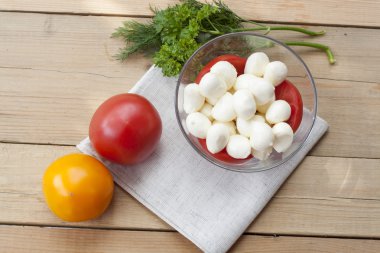Mozzarella cheese in a glass bowl, tomatoes, sliced tomatoes and herbs on a wooden table, selective focus