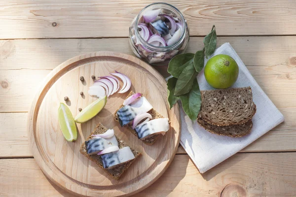 Slices of marinated mackerel with onion in a jar, lime, laurel and bread on wooden board, selective focus — Stock Photo, Image