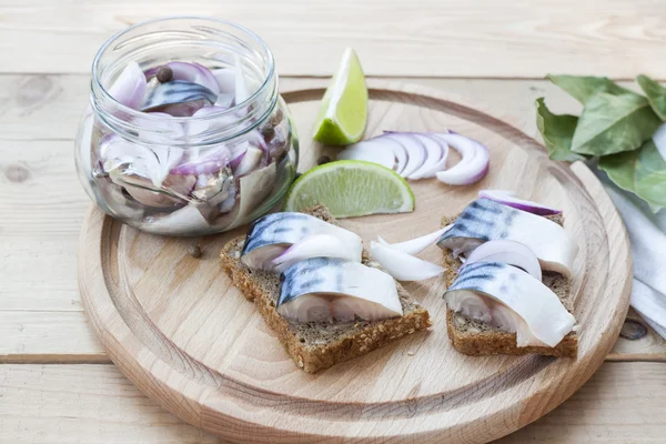 Slices of marinated mackerel with onion in a jar, lime, laurel and bread on wooden board, selective focus — Stok fotoğraf