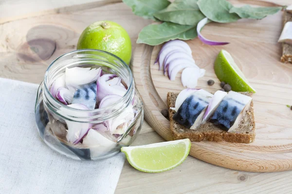 Slices of marinated mackerel with onion in a jar, lime, laurel and bread on wooden board, selective focus — Stock Fotó