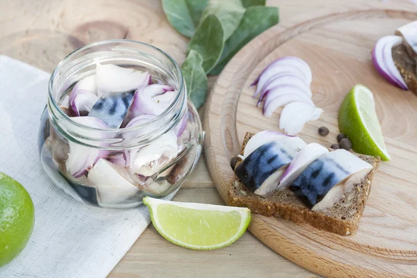 Slices of marinated mackerel with onion in a jar, lime, laurel and bread on wooden board, selective focus — Stock Fotó