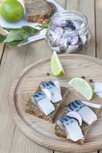 Slices of marinated mackerel with onion in a jar, lime, laurel and bread on wooden board, selective focus — Stock Photo, Image