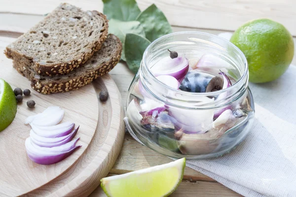 Slices of marinated mackerel with onion in a jar, lime, laurel and bread on wooden board, selective focus — Stock Photo, Image