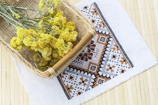 Dwarf everlast flowers bouquet in a wicker basket and napkin with embroidery on light wooden table, selective focus — Stockfoto