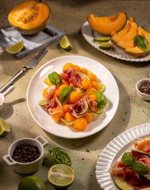 Cantaloupe melon balls paired with thin prosciutto slices, lime wedges and basil on a textured concrete surface, vibrant summer appetizer with sweet and savory contrast