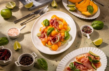 Refreshing appetizer plate featuring sweet cantaloupe melon balls, savory prosciutto, aromatic basil leaves, and zesty lime slices, preparing for a delicious summer meal