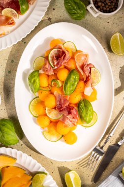Refreshing appetizer featuring vibrant orange melon balls, savory prosciutto, fresh green basil, and tangy lime slices arranged on a white plate, perfect for a light summer meal or elegant starter