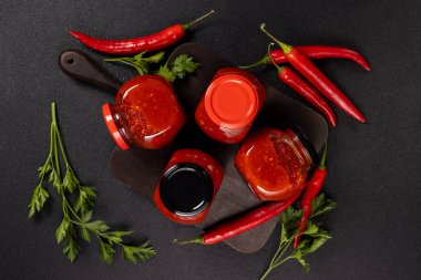 Homemade spicy sweet chili sauce in glass jars on a cutting board, surrounded by fresh red peppers and parsley, representing traditional food preparation and cooking