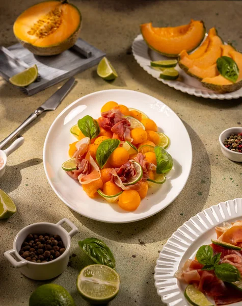Cantaloupe melon balls paired with thin prosciutto slices, lime wedges and basil on a textured concrete surface, vibrant summer appetizer with sweet and savory contrast