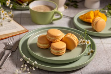 Assorted sweet macarons filling a green plate, complementing a cup of coffee and sliced mango with an open book, creating a relaxing breakfast or snack scene