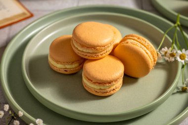 Delicate orange french macarons with a vanilla cream filling are arranged on sage green ceramic plates, creating a visually appealing dessert presentation