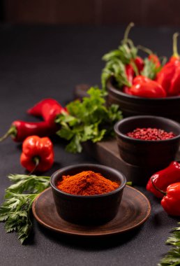 Red chili powder in a dark bowl, surrounded by fresh habanero peppers, long red chili, green herbs, and pink peppercorns, ready for use as cooking ingredients
