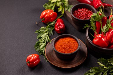 Assortment of fresh red chili peppers, habaneros, and green herbs arranged with bowls of vibrant red paprika powder and pink peppercorns on a dark background, representing spicy cooking ingredients