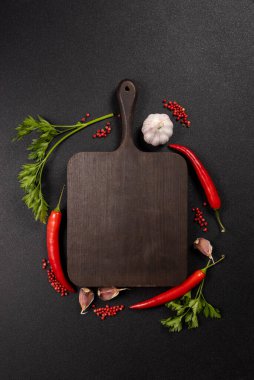 Dark, rustic wooden cutting board providing copy space, surrounded by fresh red chili peppers, garlic cloves, green parsley, and red peppercorns against a black textured background