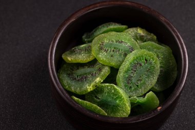 Kurumuş kivi meyve dilimleri parlak yeşil renkler ve görünür siyah tohumlar sergiliyor, dokulu bir arka planda koyu bir seramik kaseyi dolduruyor, sağlıklı ve uygun bir atıştırmalığı temsil ediyor.