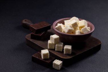 Beyaz küp şeklinde fındıklı gofretler bir kabı dolduruyor ve siyah çikolata çubuklarıyla ahşap bir kesme tahtasına saçılmış, zıt tatlı, durgun bir yaşam kompozisyonu oluşturuyor.