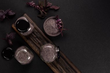 Siyah kapaklı ve seramik kâseli cam kavanozlarda mor fesleğen, koyu ahşap tahtada taze fesleğen filizi ve kırsal mutfak baharatı için siyah yüzey.