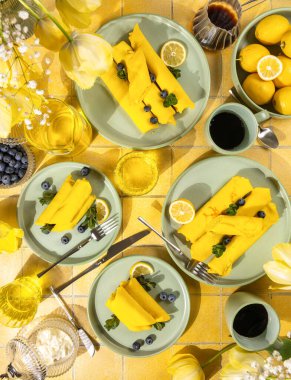 Yeşil tabaklar üzerinde yaban mersini ve nane şekeri, limon dilimleri, kahve ve sarı lalelerle kaplı parlak sarı krep yatağı.
