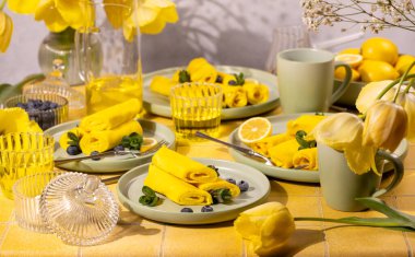 Yuvarlak sarı kreplerin yer aldığı kahvaltı masası taze yaban mersini ve nane ile süslenmiş, güneşli bir günde limon kaseleri ve çiçek açan sarı lalelerle tamamlanmış.