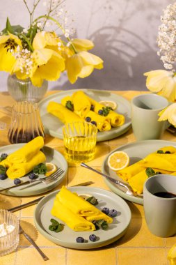 Tatlı sarı krepler sarılmış ve taze yaban mersinleri, nane yaprakları ve limon dilimleriyle süslenmiş, kahve ve meyve suyu ile servis edilmiş parlak sarı fayanslı bir masada, neşeli bir sabah kahvaltısını kutluyorlar.