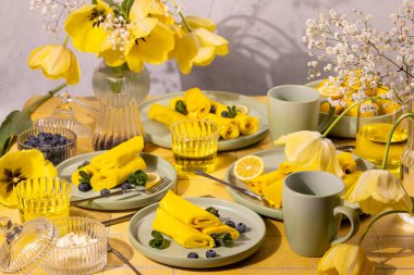 Taze yaban mersinli sarı krepler, limon dilimleri ve nane yaprakları yeşil tabaklar üzerinde düzenlenerek şenlikli bir bahar kahvaltısı için canlı sarı lalelerle süslenmiş bir masa oluşturulur.