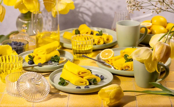 Yuvarlak sarı kreplerin yer aldığı kahvaltı masası taze yaban mersini ve nane ile süslenmiş, güneşli bir günde limon kaseleri ve çiçek açan sarı lalelerle tamamlanmış.