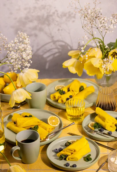 Yaban mersinli sarı krepler, limon dilimleri ve nane yaprakları açık yeşil tabaklarda servis edilir, kahve, meyve suyu ve yumuşak sabah ışığında taze lalelerle bahar brunch 'ının keyfini çıkarırlar.