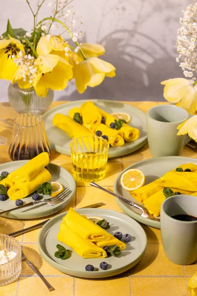 Tatlı sarı krepler sarılmış ve taze yaban mersinleri, nane yaprakları ve limon dilimleriyle süslenmiş, kahve ve meyve suyu ile servis edilmiş parlak sarı fayanslı bir masada, neşeli bir sabah kahvaltısını kutluyorlar.