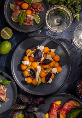 İçinde kavun topu salatası, feta, mor fesleğen ve limon bulunan aperatif tabaklar, yanında jambona sarılı kavun, koyu bir masada iki bardak beyaz şarapla servis edilir.
