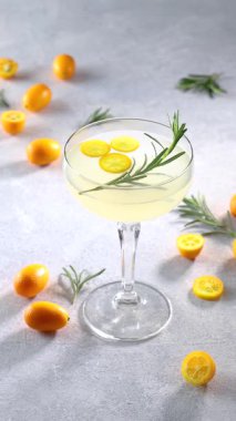 Ağır çekimde dönen kumkatlı narenciye kokteyli ve taze bir biberiye filizi. Açık gri arka planda yarı yarıya meyveler.