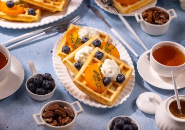 Taze şeftali, yaban mersini ve kremalı waffle sıcak çay, ceviz ve taze yaban mersinli beyaz bir tabakta servis edilir ve tatlı ve doyurucu bir kahvaltı masası oluşturur.