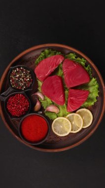 Limon dilimleri, sarımsak ve biber gibi çeşitli baharatlarla taze çiğ ton balığı filetoları koyu bir zemin üzerinde dönen kırmızı biber tabakları sağlıklı omega 3 zengin besinleri temsil ediyor.