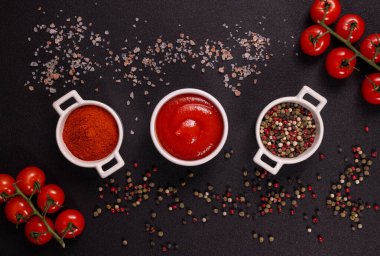 Bir kase kırmızı biber tozu, domates sosu ve karışık biberler taze kiraz domatesleri ve koyu bir masanın üzerine serpiştirilmiş deniz tuzu ile çevrilidir.