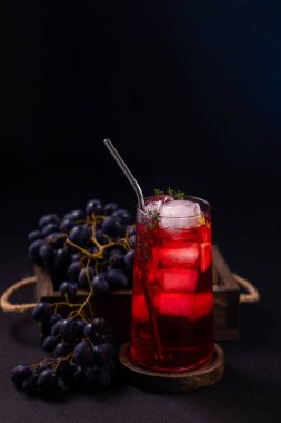 Buz ve metal pipetle soğutulmuş kırmızı kokteyl, taze kekik ile süslenmiş, kırsal ahşap bir sandıkta koyu olgun üzümlerle eşleştirilmiş, minimal siyah bir stüdyo arka planında.