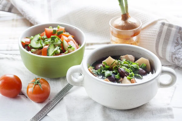 Vegetable soup with beans and chicken, fresh herbs in a clay pot, salad of tomatoes and cucumbers with greens in a green bowl on a white wooden table. — Stock Image Vegetable soup with beans and chicken, fresh herbs in a clay pot, salad of tomatoes and cucumbers with greens in a green bowl on a white wooden table. Stock Picture