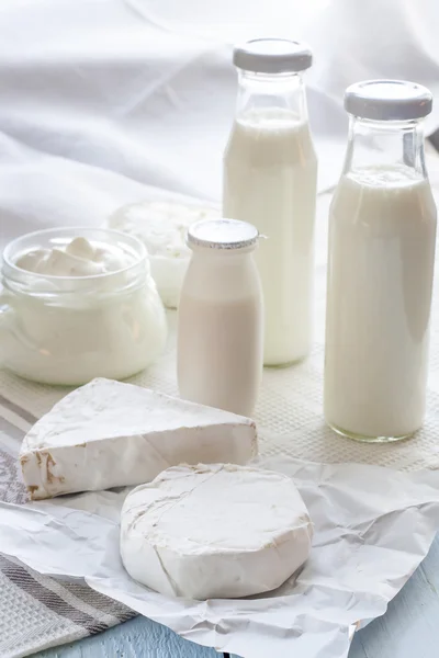 Dairy products. Milk in glass bottle, yogurt, sour milk cheese, sour cream, camembert and brie with checkered napkin on light blue wooden table — Stock Fotó