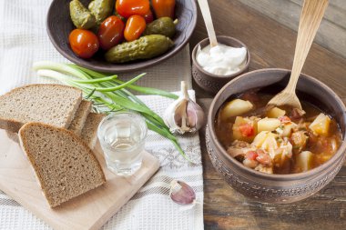 Traditional ukrainian vegetable soup - borsch, marinated tomatoes and cucumbers, sour cream, sliced bread, herbs and garlic, vodka in glass cup at dark wooden table.
