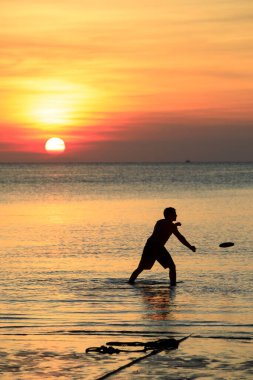 Adam güzel günbatımına karşı plajda disk uçuruyor.