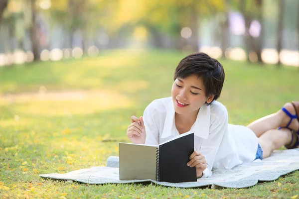 Jonge vrouw liggen op groen gras park met potlood en Opmerking boek — Stockfoto