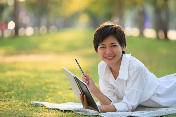 Młoda kobieta leżący na zielonej trawie parku z ołówkiem i Uwaga książki — Zdjęcie stockowe