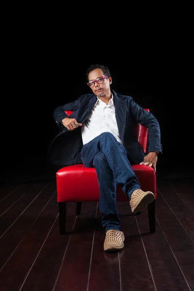 Portrait of 45 years old asian man sitting on red sofa against b — Stock Photo, Image