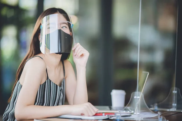 young Asian business woman working in cafe with medical face mask and table shield protection, social distancing of the new normal lifestyle after epidemic of coronavirus COVID-19