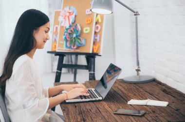 Asyalı bir kız evde dizüstü bilgisayarla çalışıyor. Evde oturuyor, arıyor ve not yazıyor.