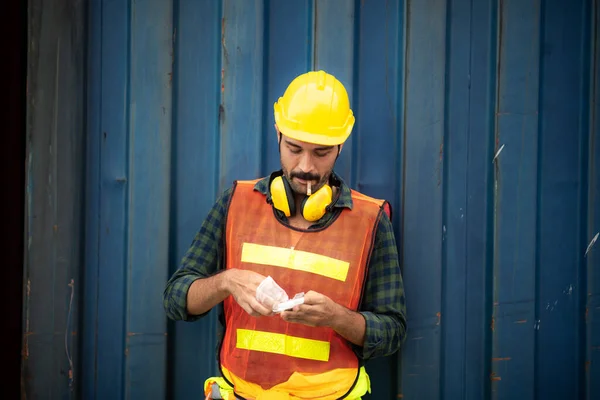 an employee foreman men worker smoking cigarette tobacco nicotine whlie take a break, relax work lifestyle concept, container site, outdoor smoking, men wearing safety vest, helmet and headphone, unhealthy concept