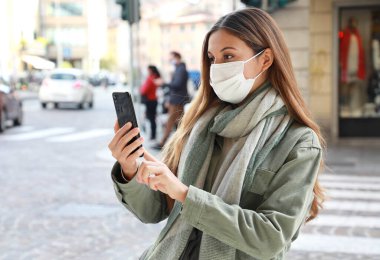Happy young woman in surgical mask for protection against the virus stands on city street makes video call clipart