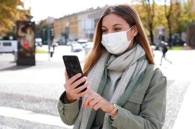 Şehirde yüz maskesi takan akıllı telefon kullanan genç bir kadının portresi.